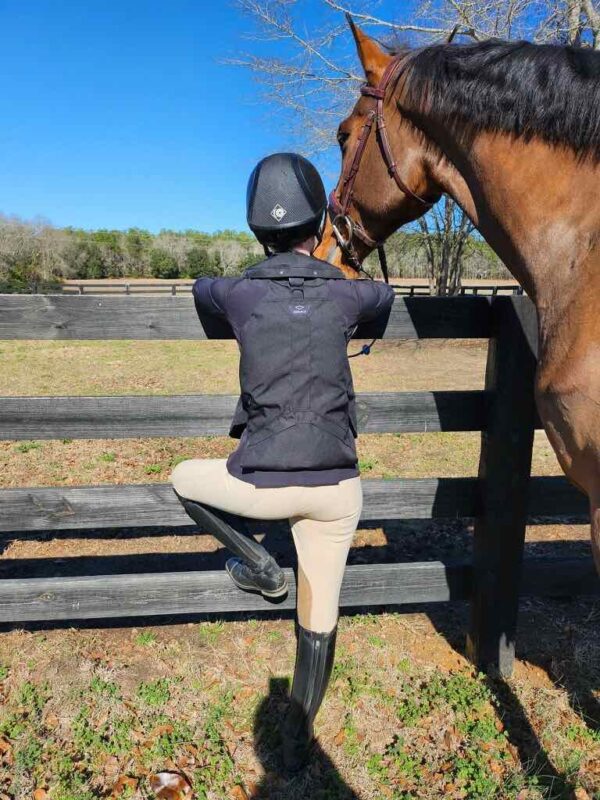 A person in riding gear kneels down to pet a horse.